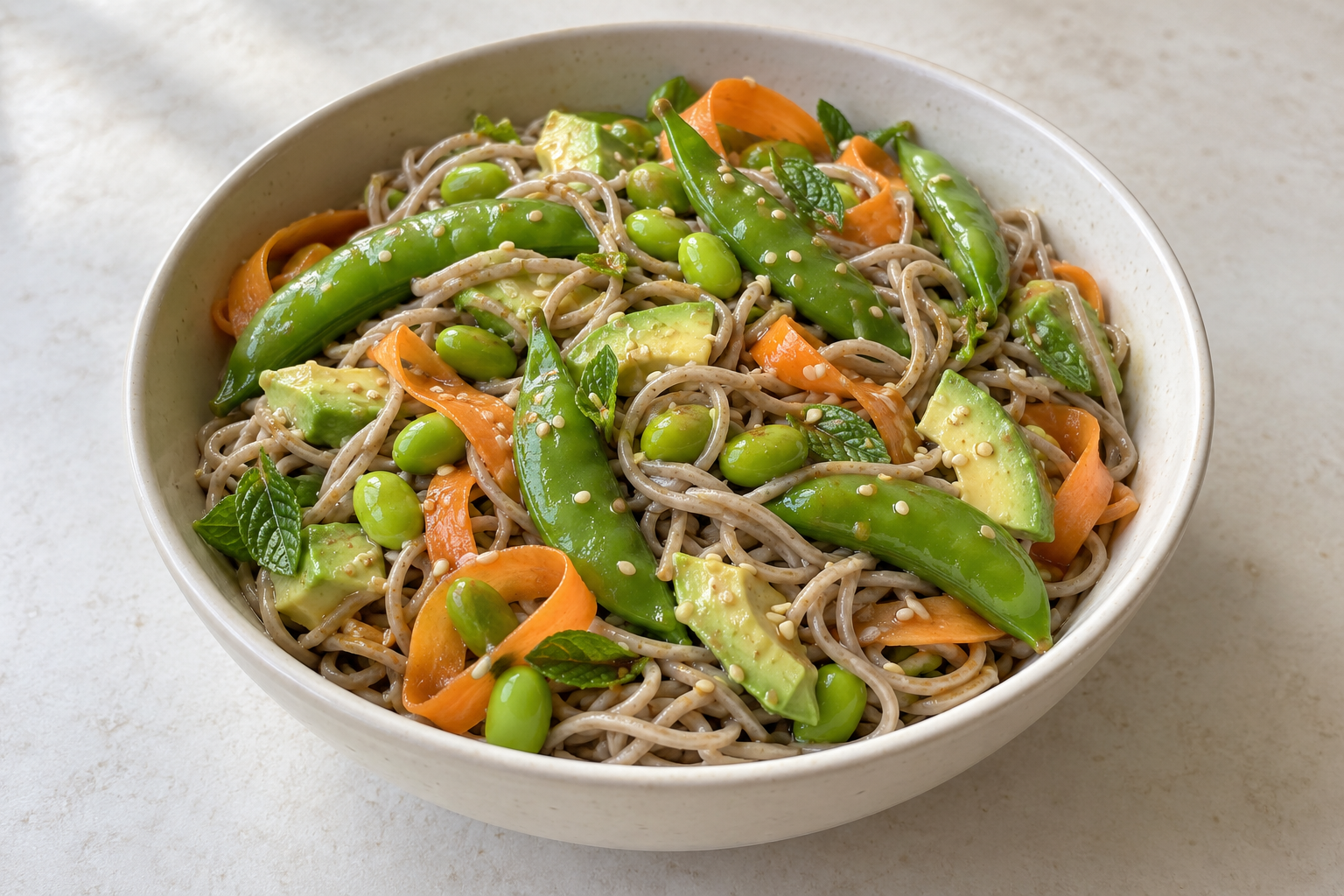 Sesame soba noodles with plump snap-pea pods, shelled edamame, avocado, carrot ribbons, mint and sesame seeds in a bowl.